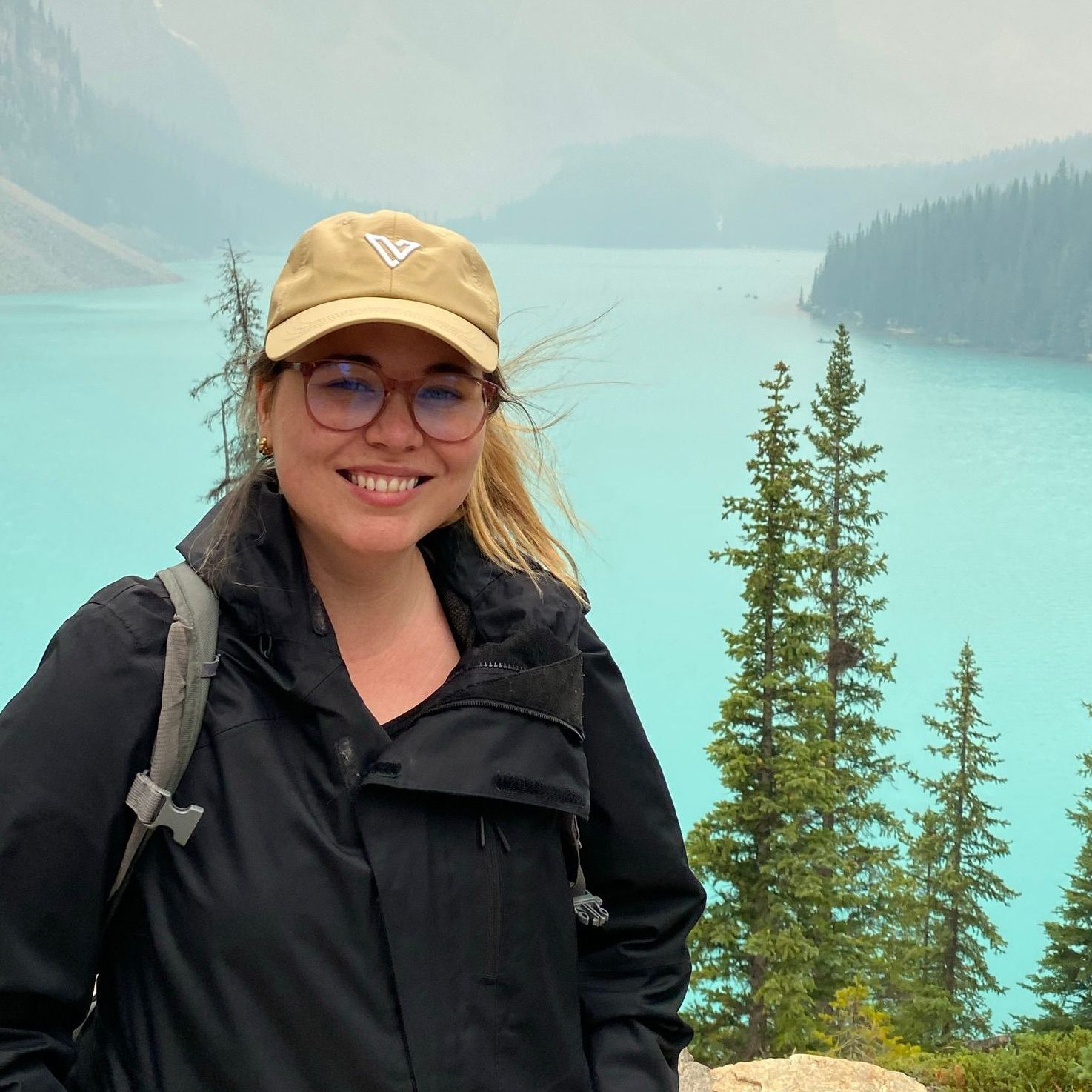 Headshot of Jaida Regan in front of Lake Louise wearing a black jacket and cream hat with glasses.