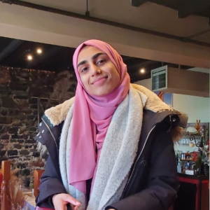 Headshot of Amal Abdullah wearing a dark grey parka and a pink hijab with a brick wall background.